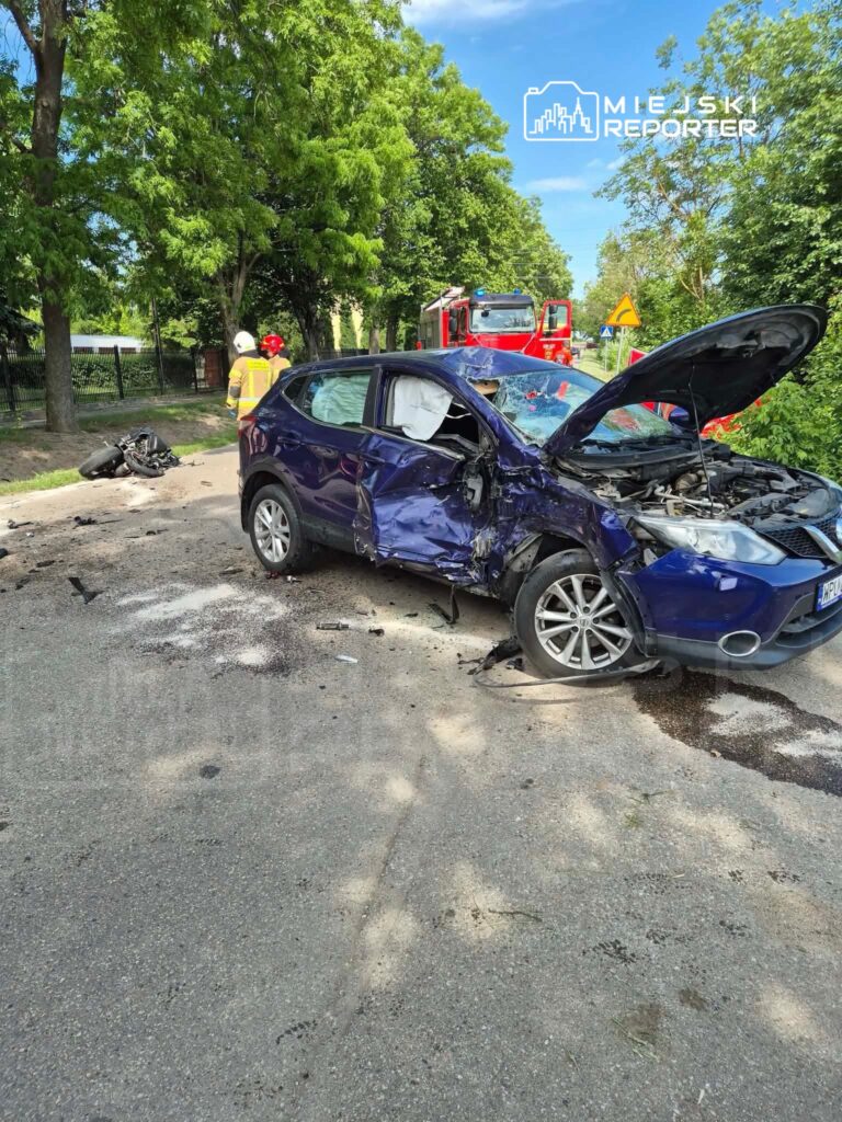 Rozbity niebieski samochód osobowy z uszkodzoną karoserią stoi na drodze, obok strażacy i wrak motocykla.