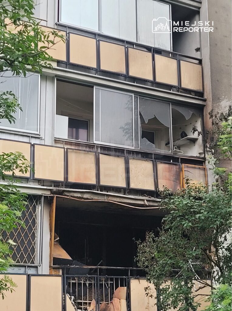 Zniszczona elewacja budynku mieszkalnego z wybitymi oknami i uszkodzoną balustradą. W tle widoczna zieleń.