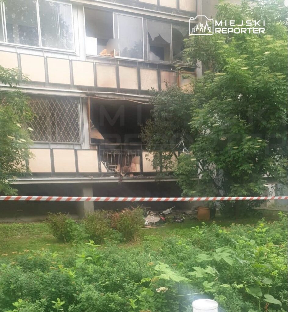 Uszkodzone okna mieszkania na pierwszym piętrze bloku, z widocznymi zniszczeniami wewnętrznymi, otoczone roślinnością.