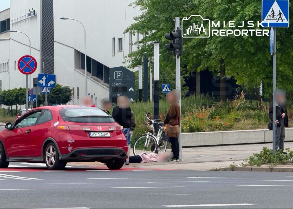 Czerwony samochód zatrzymany na pasie drogowym, obok grupa osób, z rowerem i leżącą osobą na chodniku.