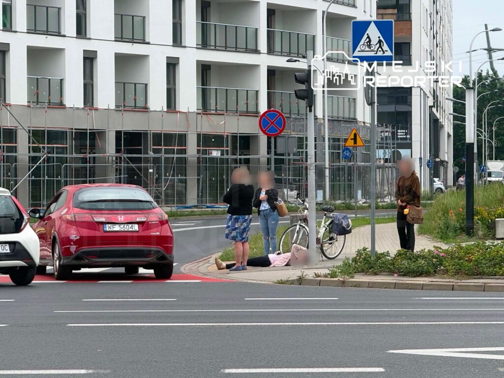 Grupa osób stoi obok leżącej osoby na chodniku, w tle nowoczesny budynek z przeszkloną fasadą.