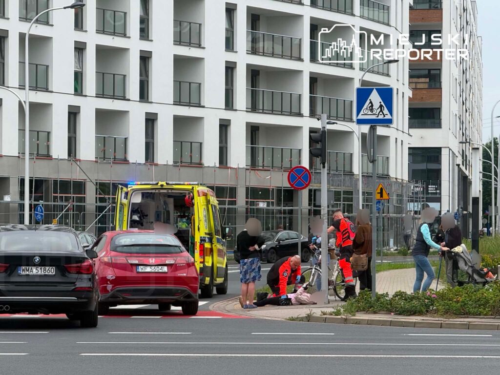 Ratownicy medyczni udzielają pomocy osobie leżącej na chodniku obok nowoczesnego budynku mieszkalnego.