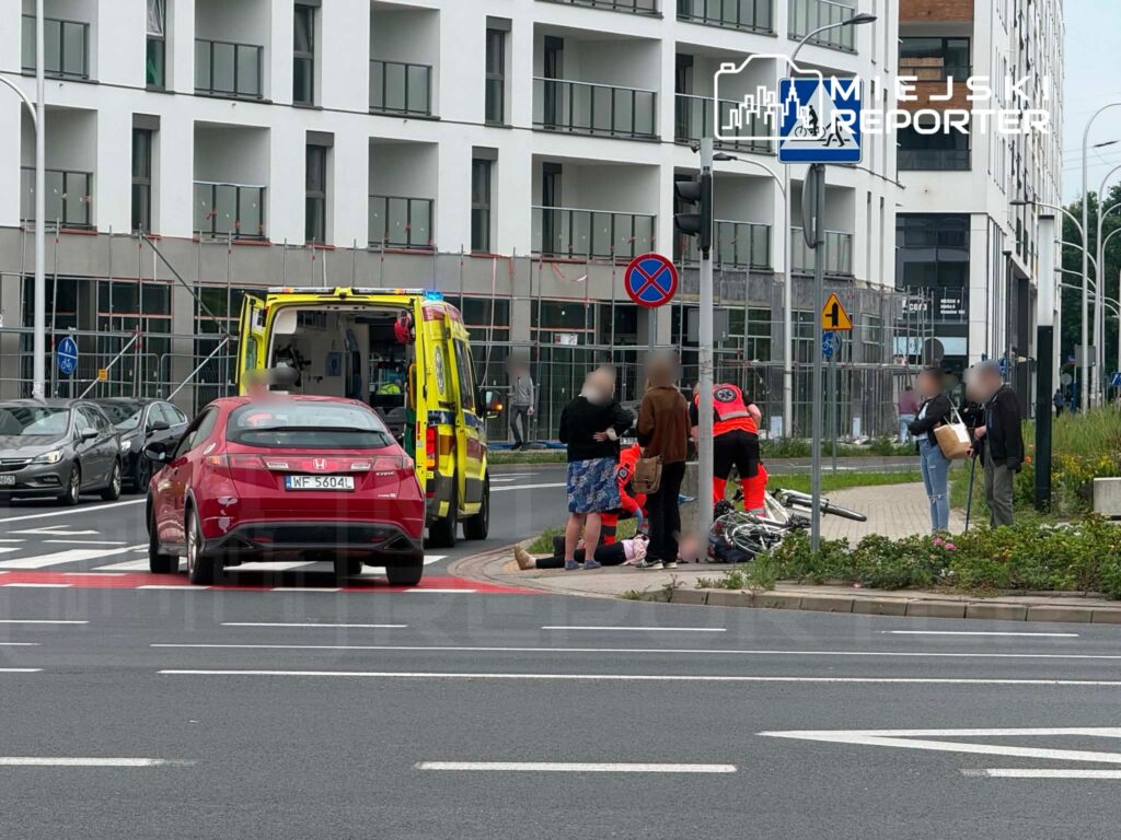 Na chodniku przy nowoczesnym budynku grupa osób otacza leżącego rowerzystę, a obok zatrzymuje się karetka.