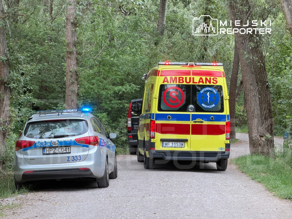 Radiowóz policyjny i karetka pogotowia zaparkowane na leśnej drodze, otoczone gęstą roślinnością.