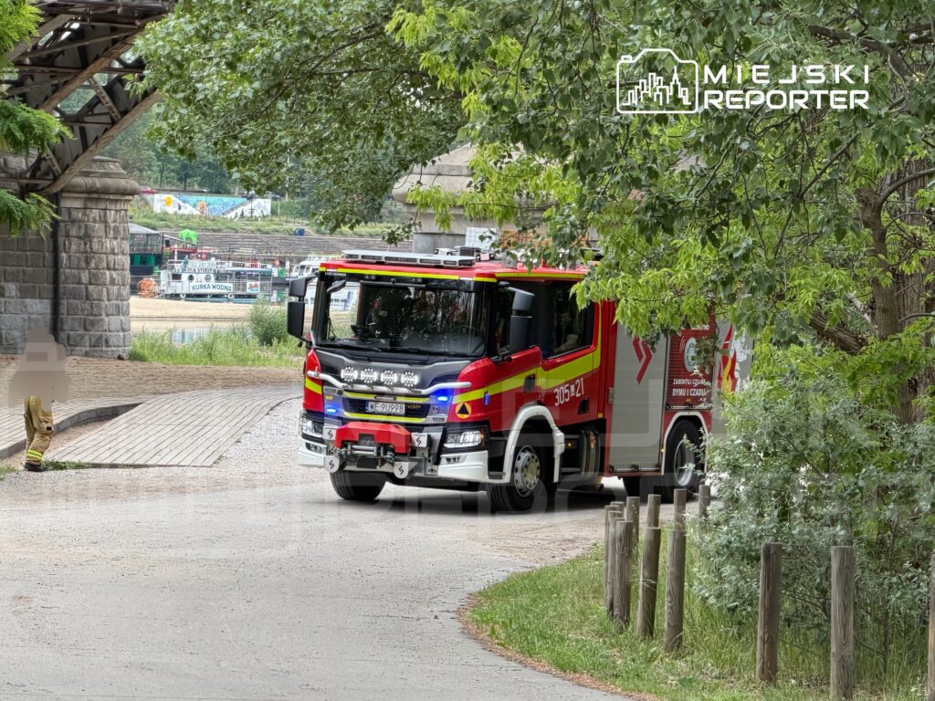 Wóz strażacki zatrzymany na ścieżce w pobliżu wody, otoczony zielonymi drzewami i drewnianymi słupkami.