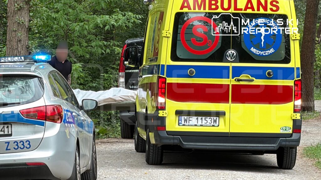 Policjant przy radiowozie obserwuje ratowników medycznych przenoszących przykryte ciało w lesie.