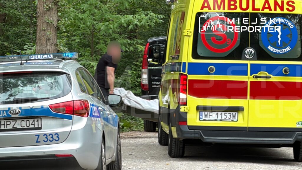 Mężczyzna w czarnym ubraniu przenosi przykryty obiekt w kierunku karetki w lesistym otoczeniu.