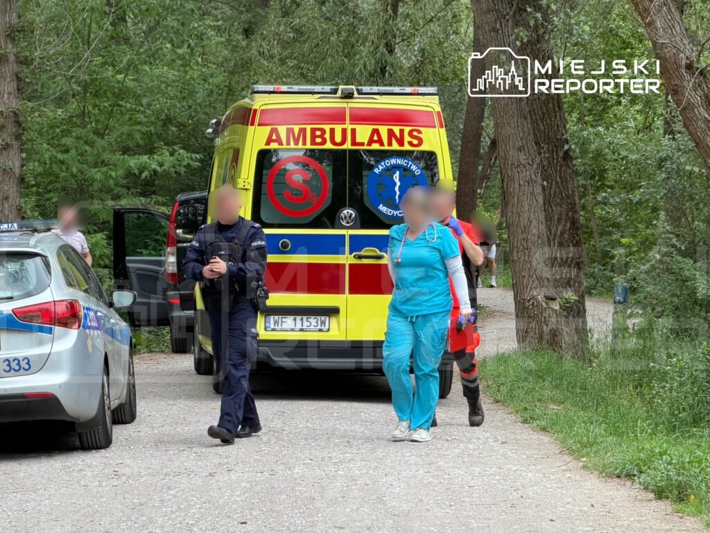 Ratownik medyczny w niebieskim uniformie prowadzi osobę w kierunku karetki, obok stoi policjant przy radiowozie.