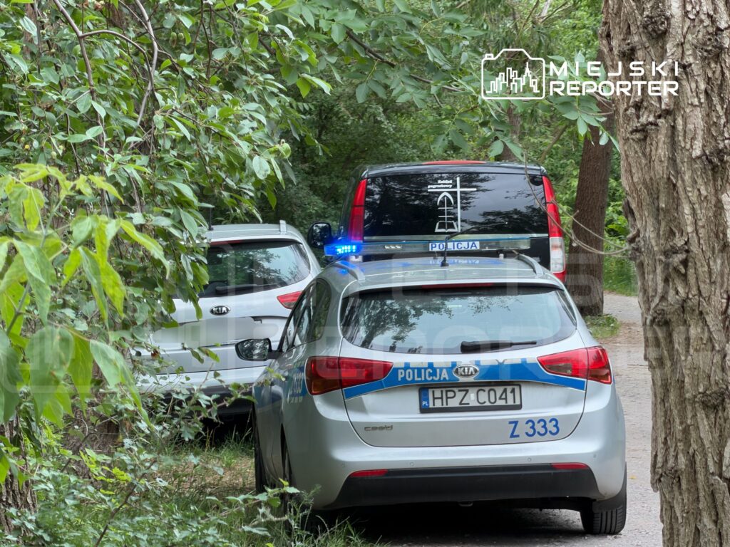 Na leśnej drodze zaparkowane są dwa samochody, w tym radiowóz z włączonymi sygnałami świetlnymi.