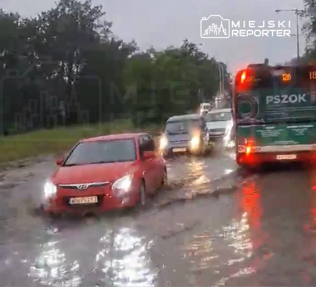 Czerwony samochód osobowy porusza się po zalanej ulicy, obok przejeżdża autobus komunikacji miejskiej.