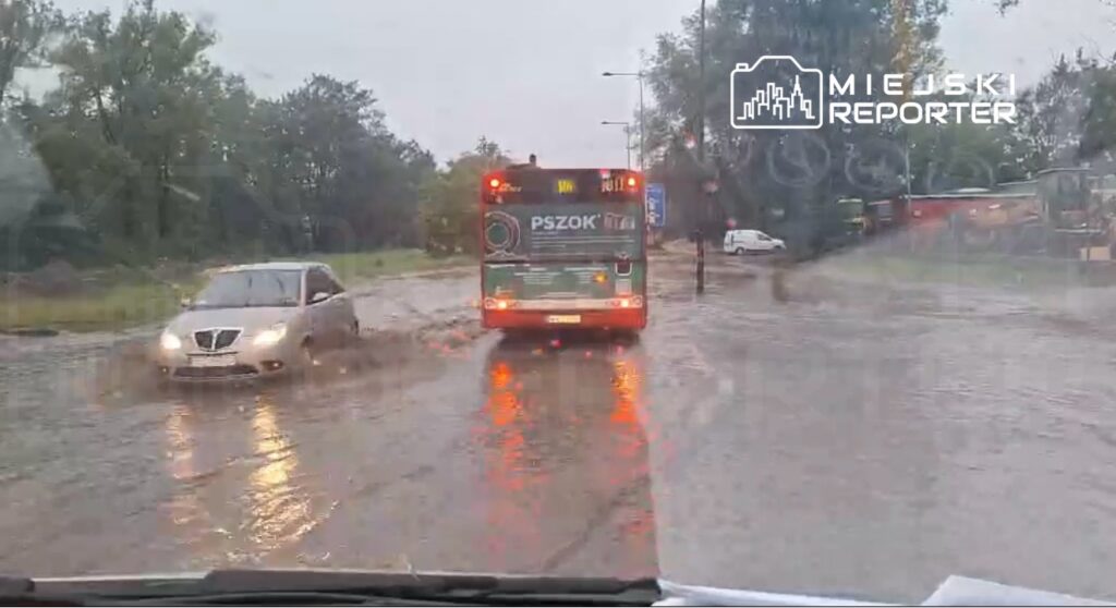 Zatopiona ulica z samochodem osobowym oraz miejskim autobusem poruszającym się w wodzie.
