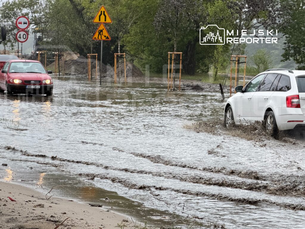 Dwa samochody poruszają się po zalanej drodze, obok znaków drogowych i budowy.