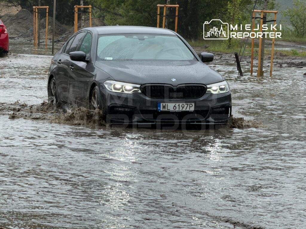 Czarny samochód osobowy porusza się przez zalaną ulicę, woda sięga do połowy kół. W tle widać inne pojazdy.