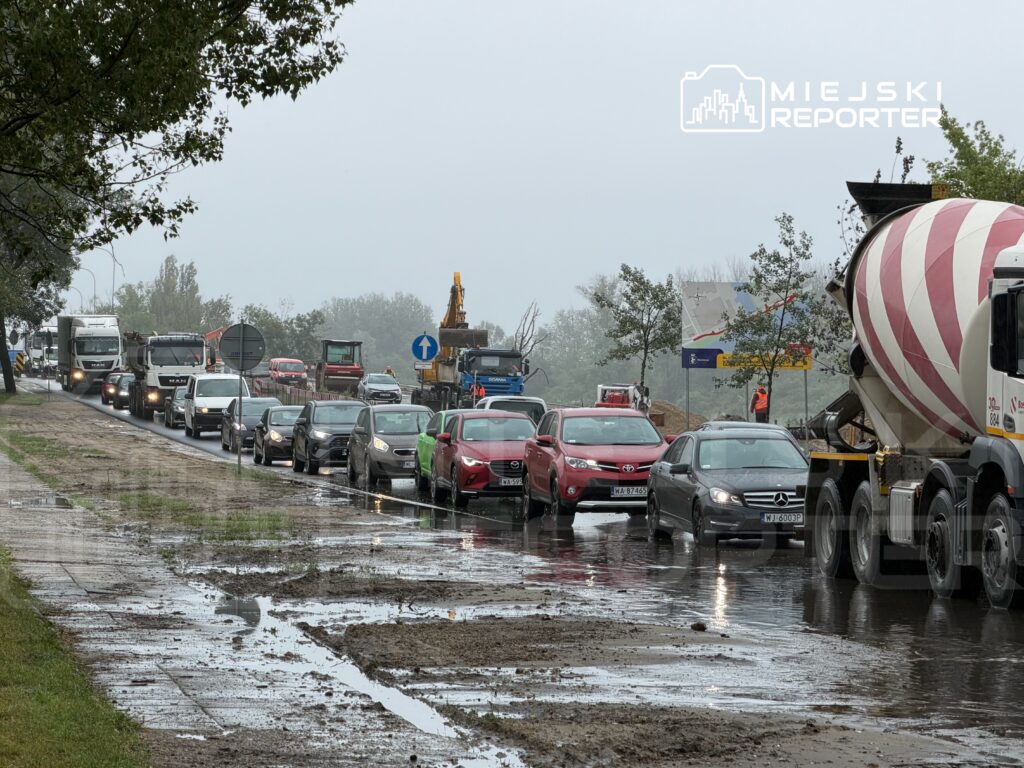 Ruch samochodowy w deszczowy dzień na drodze z budową, z widocznymi ciężarówkami i samochodami osobowymi.