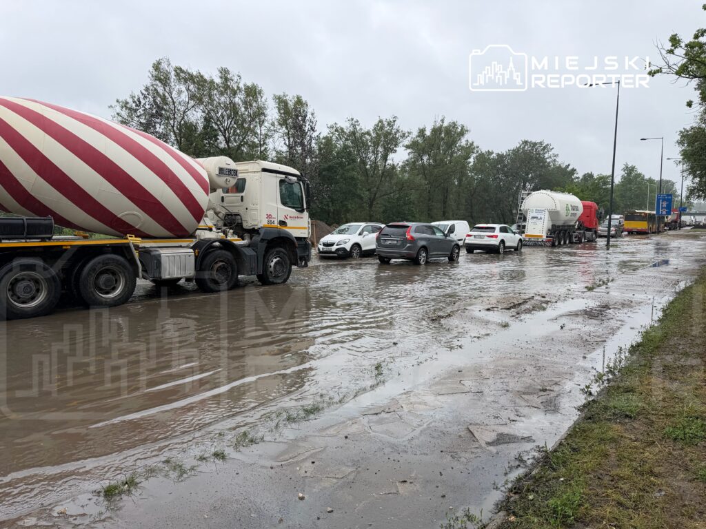 Ruch samochodowy na zalanej drodze, z betonowymi pojazdami i osobowymi autami w deszczowej aurze.