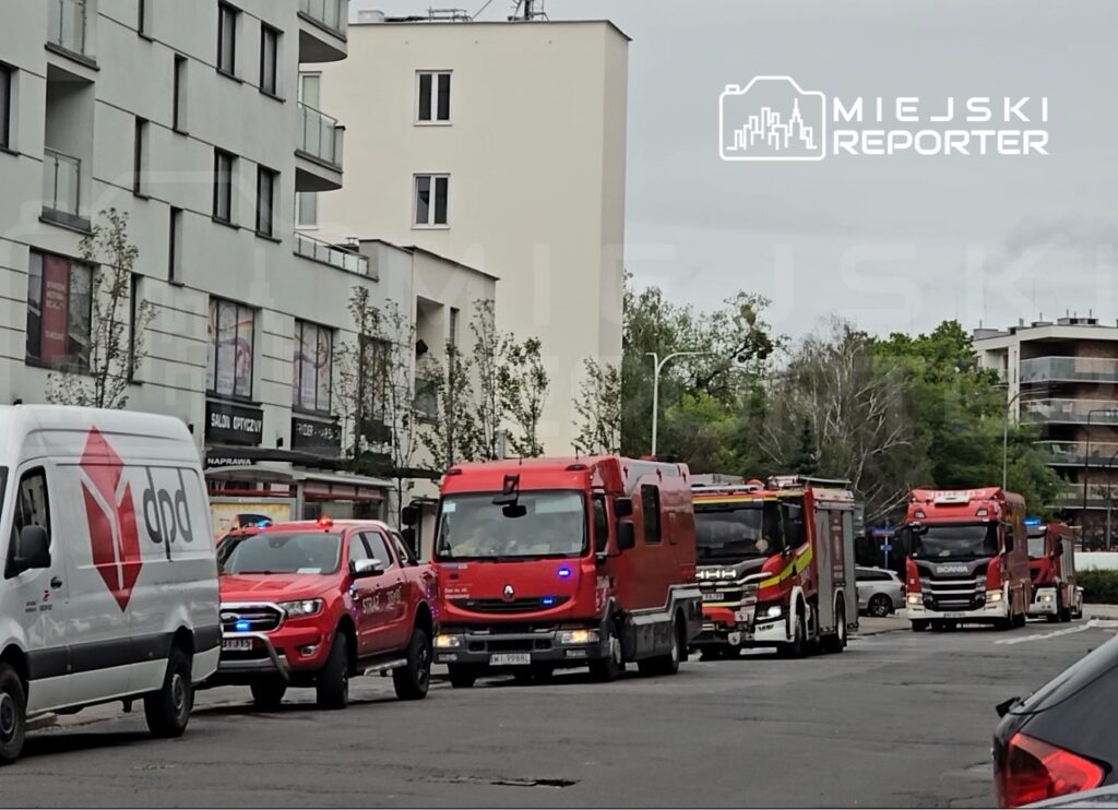 Rząd samochodów straży pożarnej oraz pojazdów służb ratunkowych zaparkowanych na ulicy w miejskim otoczeniu.