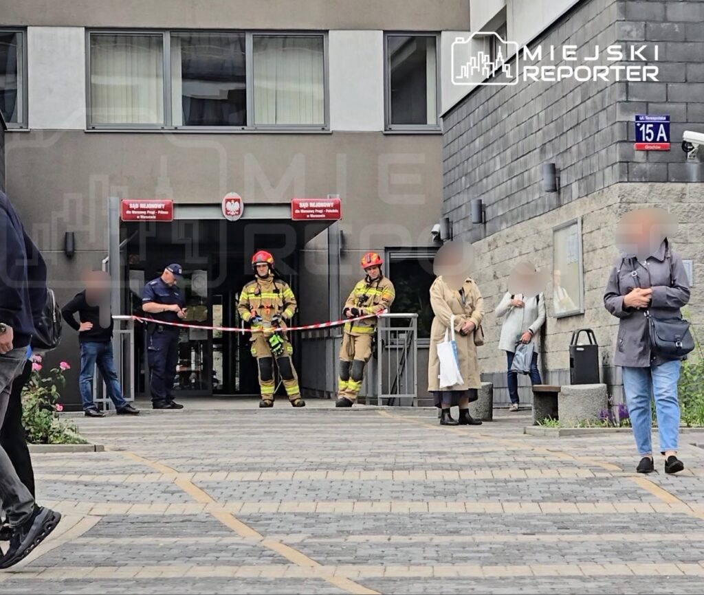 Grupa strażaków w mundurach i kaskach zabezpiecza wejście do budynku, podczas gdy przechodnie obserwują sytuację.