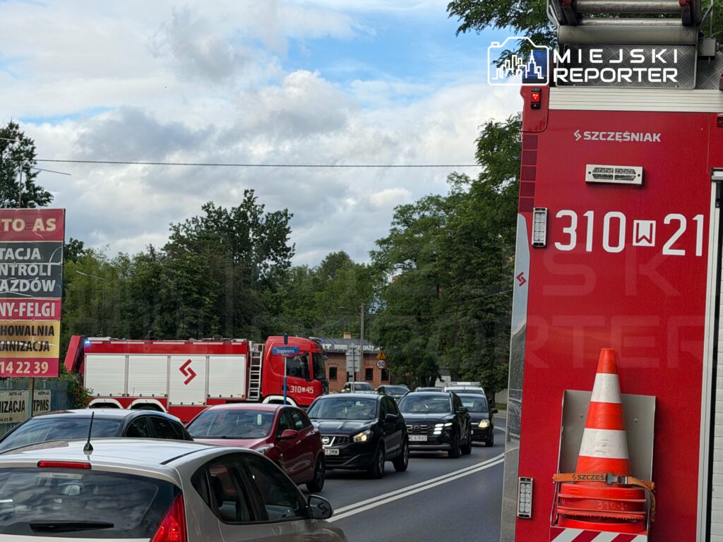 Czerwony wóz strażacki zatrzymany na ruchliwej drodze, obok znak informacyjny oraz stojące w korku samochody.