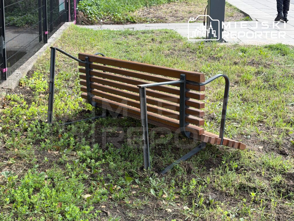 Złamana ławka parkowa z drewnianymi deskami, leżąca na trawie w otoczeniu zieleni i ogrodzenia.