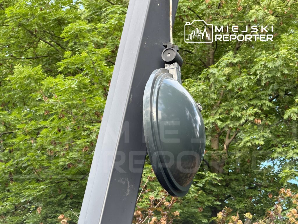 Czarna lampa uliczna zamontowana na metalowym słupie wśród zieleni drzew.