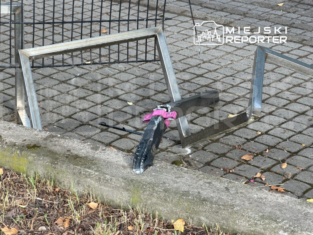 Złamany metalowy stelaż leżący na kostce brukowej, obok widoczny kawałek materiału związany różową taśmą.