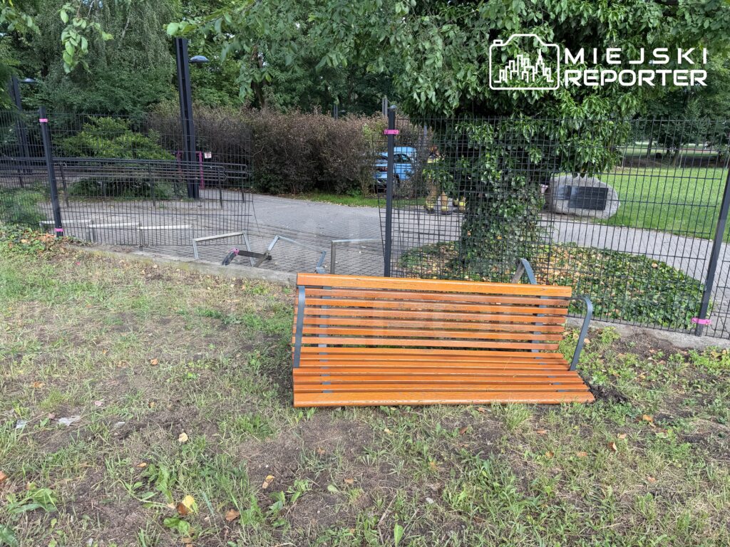 Ławka z drewna w parku, umieszczona na trawie, z widoczną ogrodzeniem i alejką w tle.