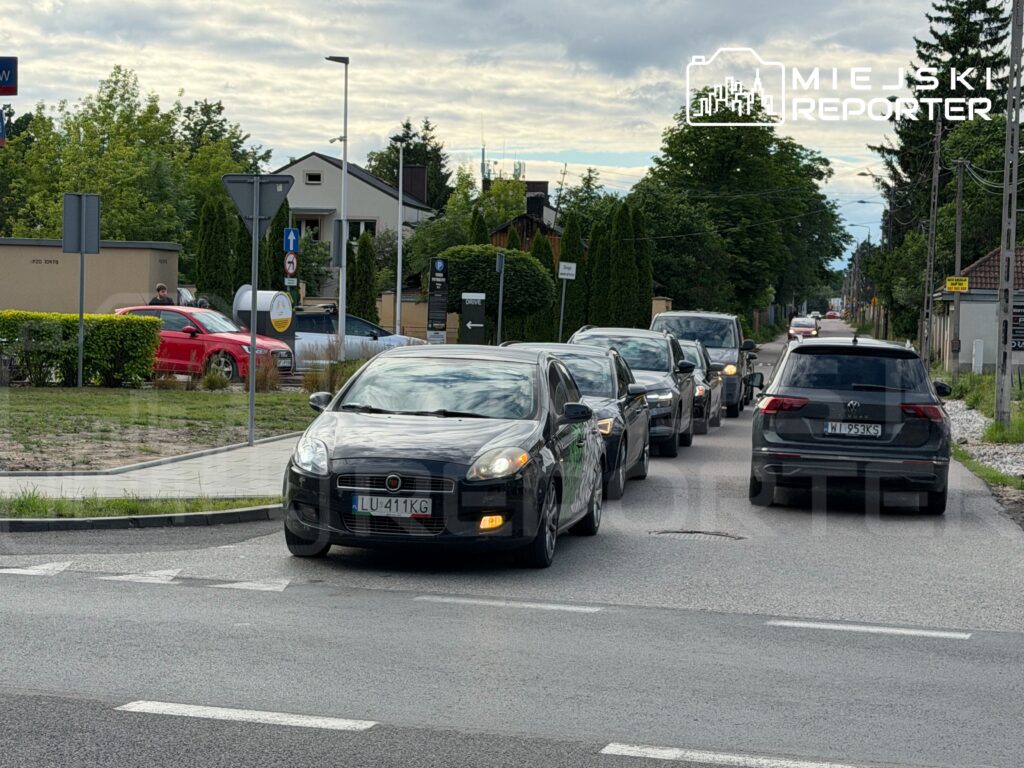 Ruch samochodowy na ulicy z kilkoma pojazdami w kolejce, otoczony zielenią i znakami drogowymi.