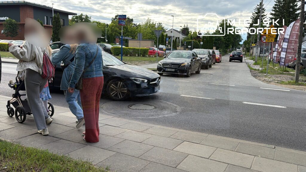 Grupa osób na chodniku rozmawia obok jezdni, w tle stoi czarny samochód i budynek McDonald's.