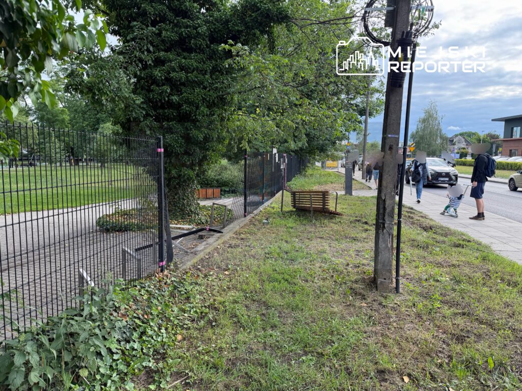 Grupa ludzi spacerujących wzdłuż chodnika obok ogrodzenia, w tle nowoczesne budynki i zieleń.