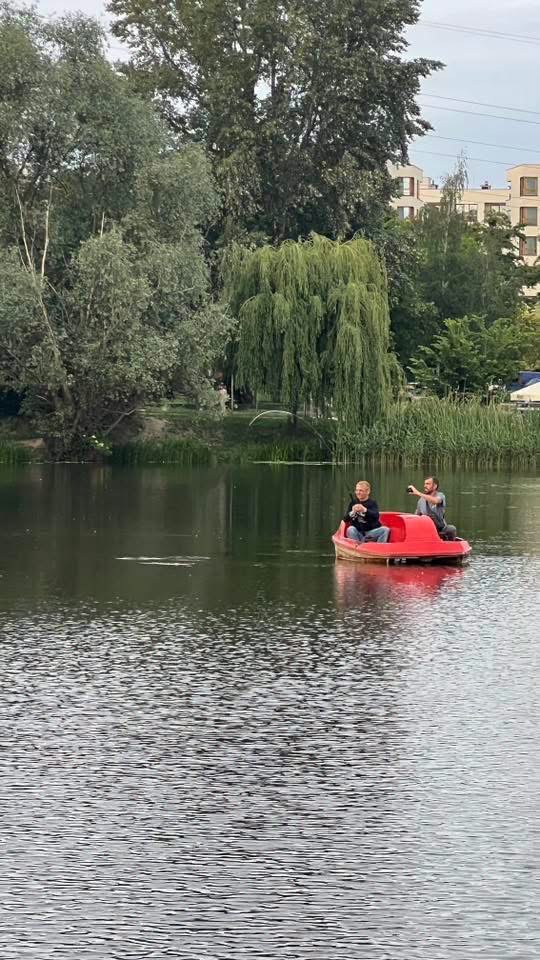 Dwoje ludzi na czerwonym rowerze wodnym pływających po jeziorze otoczonym drzewami i budynkami.