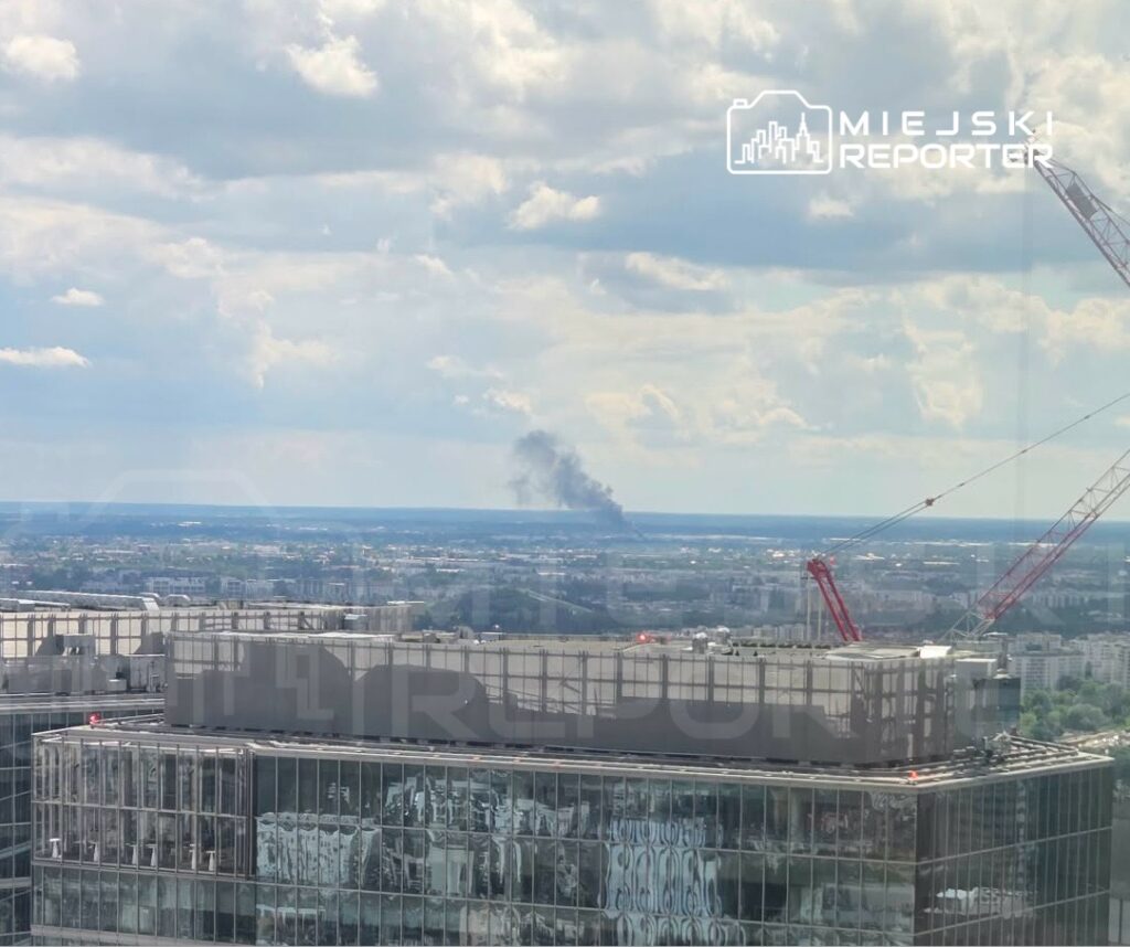 Na horyzoncie widoczny dym unoszący się nad miastem, w tle nowoczesny biurowiec z przeszkloną fasadą.