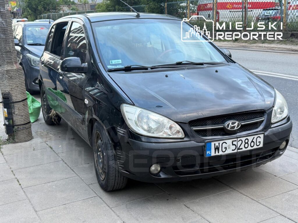 Czarny samochód osobowy zaparkowany na chodniku obok palmowego drzewa, w tle widoczne inne pojazdy.