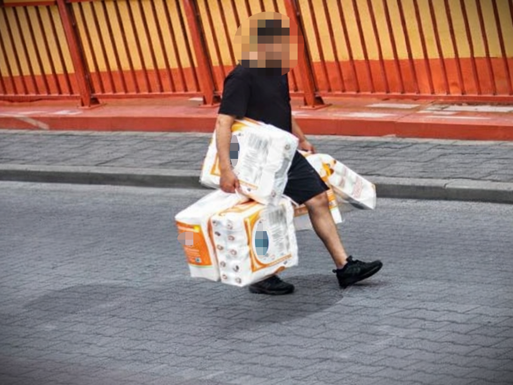 Mężczyzna niosący cztery paczki papieru toaletowego na ulicy, obok pomarańczowego ogrodzenia.