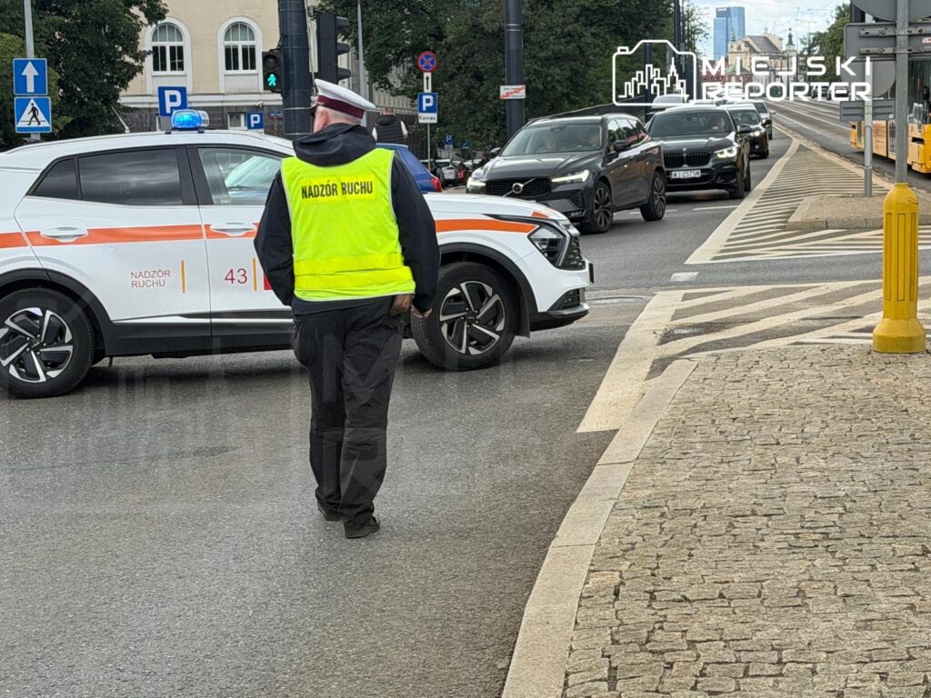 Mężczyzna w kamizelce odblaskowej nadzoruje ruch na skrzyżowaniu, obok zaparkowane samochody i sygnalizacja świetlna.