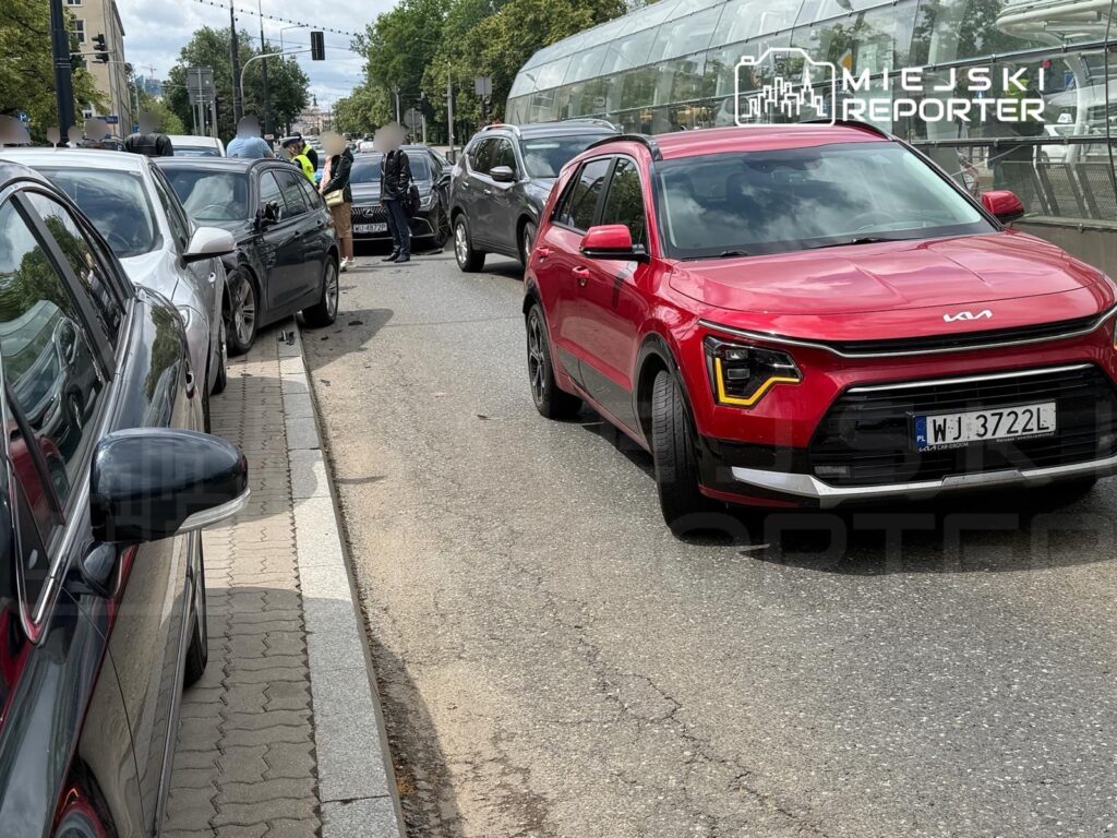 Ruch uliczny z kilkoma samochodami zaparkowanymi wzdłuż krawężnika; na pierwszym planie czerwony SUV.