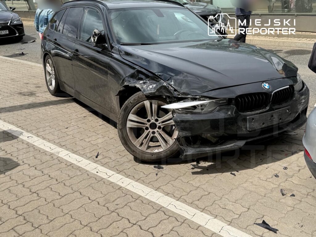 Czarny samochód osobowy marki BMW z uszkodzeniami bocznymi zaparkowany na chodniku w miejskim otoczeniu.