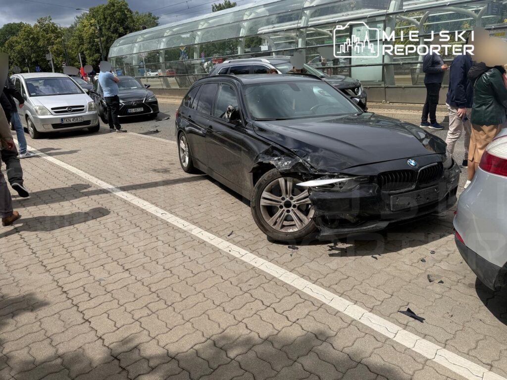 Czarny samochód osobowy z uszkodzoną przednią częścią stoi na drodze, wokół zbierają się ludzie, obserwując zdarzenie.