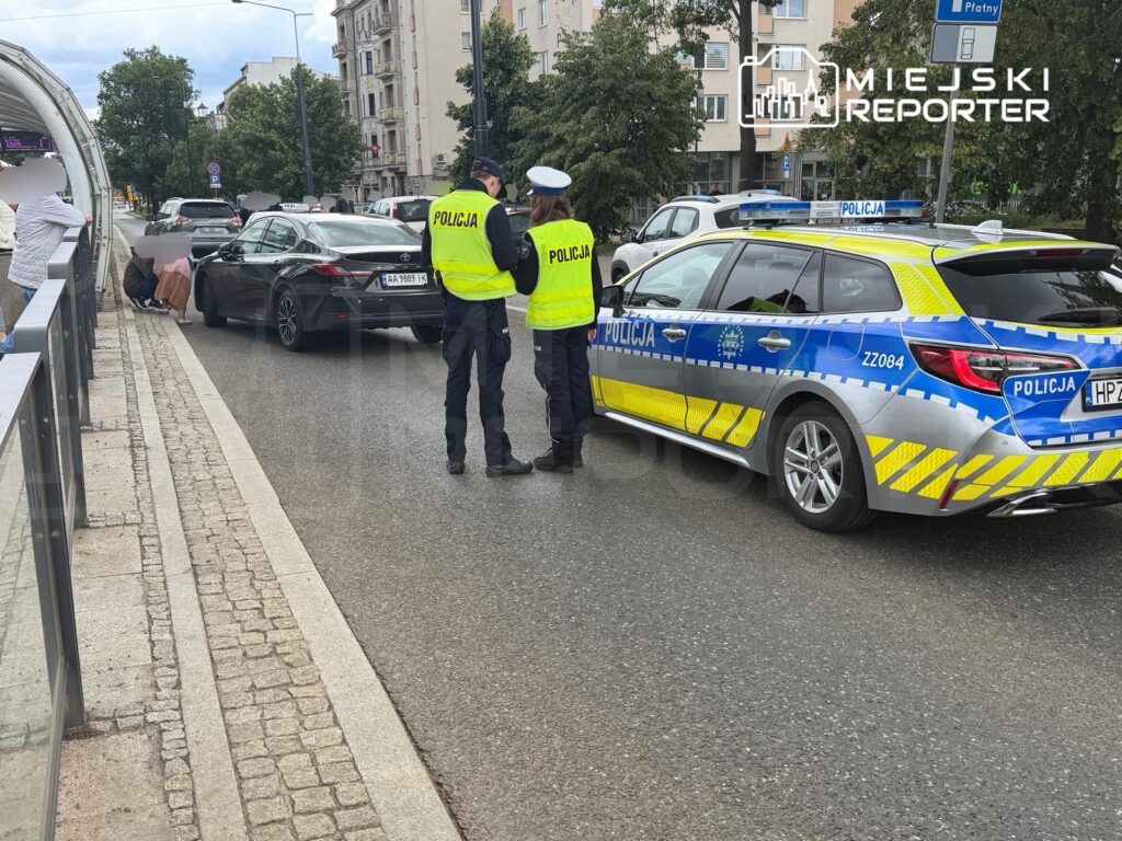 Policjanci w żółtych kamizelkach rozmawiają z kierowcą samochodu osobowego na ulicy, obok przystanku komunikacji miejskiej.