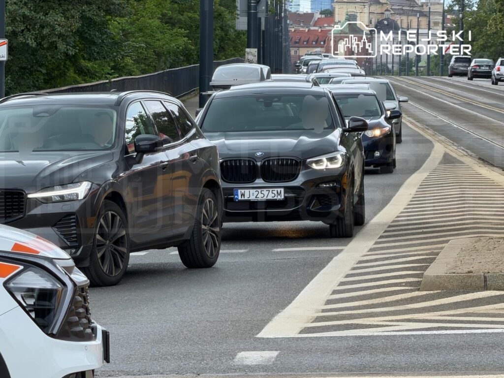 Korkujący ruch samochodowy na miejskiej ulicy, z widocznymi pojazdami osobowymi w różnych kolorach.