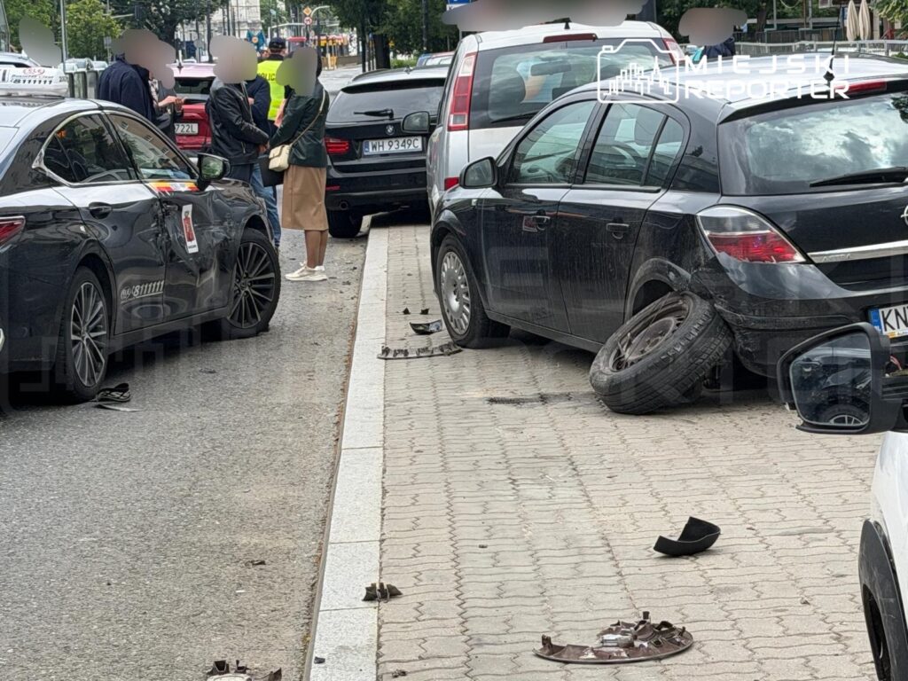 Czarny samochód osobowy z uszkodzeniami na boku stoi na chodniku, obok grupy ludzi rozmawiających z policjantem.