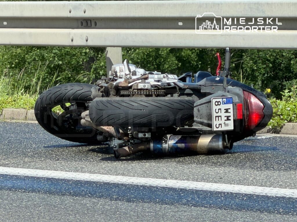 Przewrócony skuter leży na drodze obok bariery ochronnej, w otoczeniu zieleni.