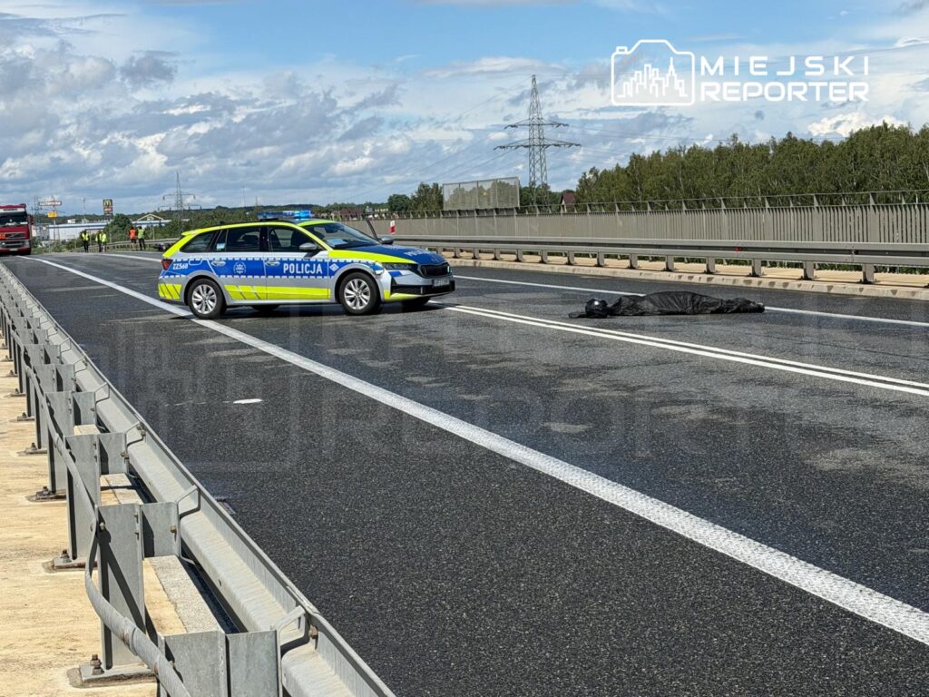 Policjant przy samochodzie służbowym zabezpiecza miejsce zdarzenia na drodze, gdzie leży przykryta sylwetka.