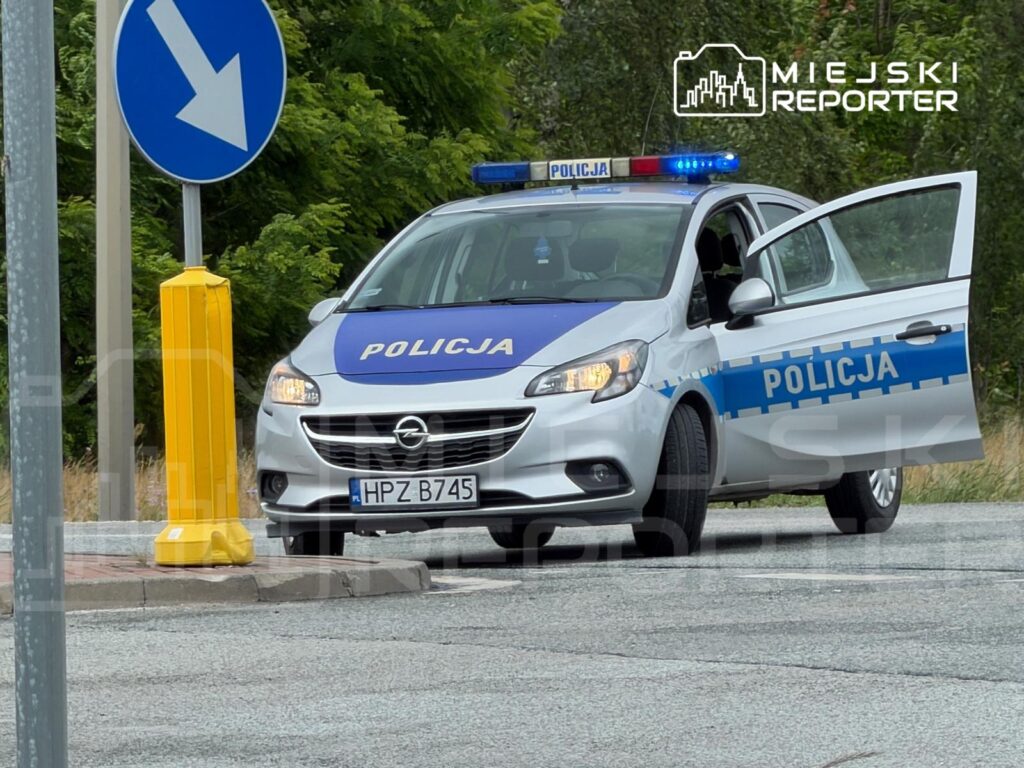 Policyjny samochód osobowy z otwartymi drzwiami zatrzymany przy znaku drogowym wskazującym kierunek.