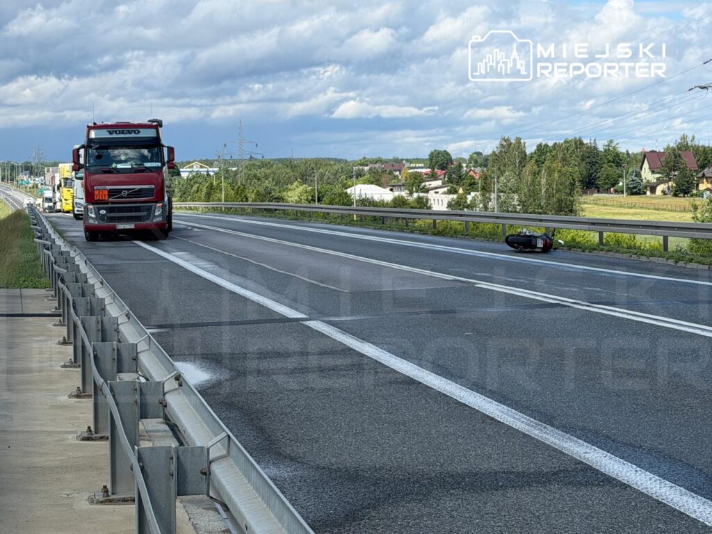 Czerwony ciężarowy samochód marki Volvo jedzie po drodze, obok leżący motocykl na poboczu. W tle zielone drzewa i zabudowania.