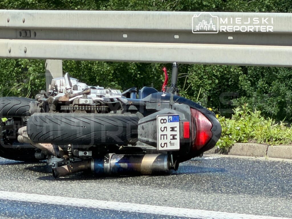 Motocykl leżący na boku na jezdni, z uszkodzoną częścią silnikową i widocznymi oponami. W tle zieleń.