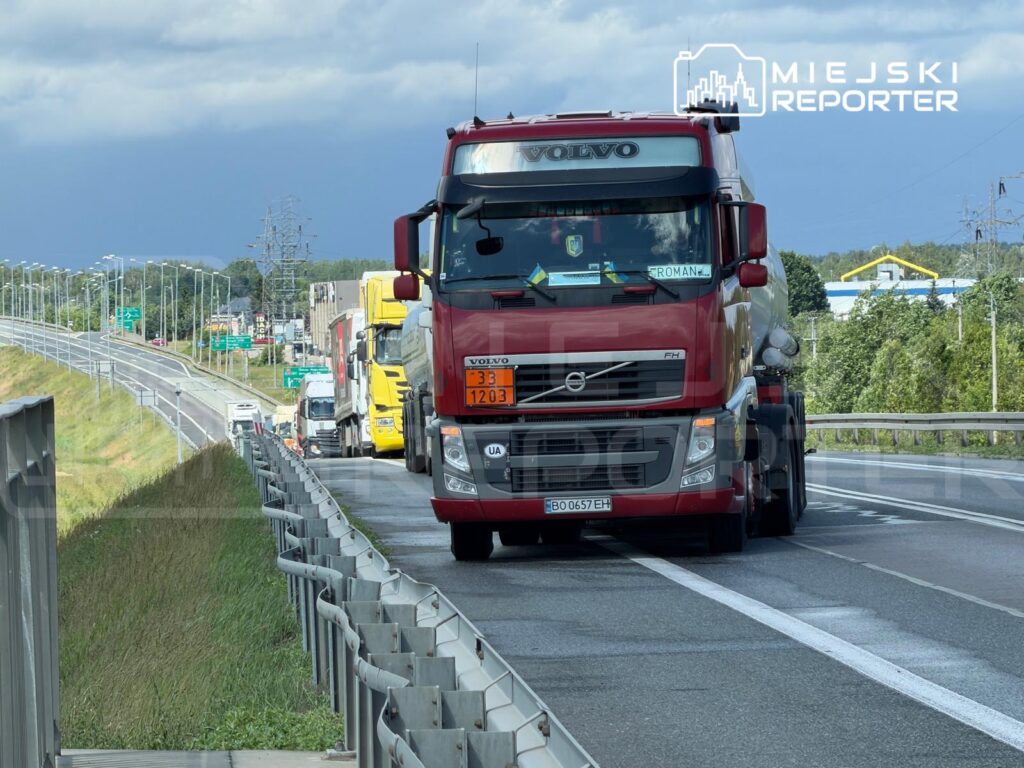 Czerwony ciężarowy pojazd marki Volvo porusza się w kierunku zachodnim, obok zatoru drogowego na autostradzie.