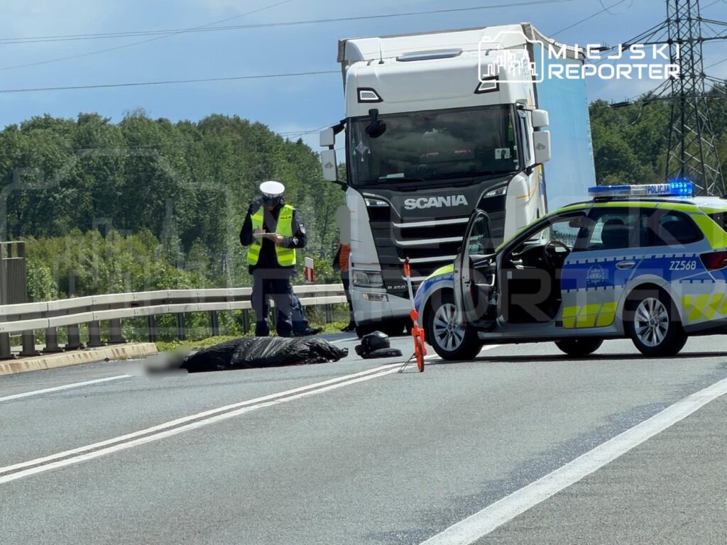 Policjant w kamizelce odblaskowej notuje zdarzenie na drodze, obok leży przykryta czarna folią sylwetka.