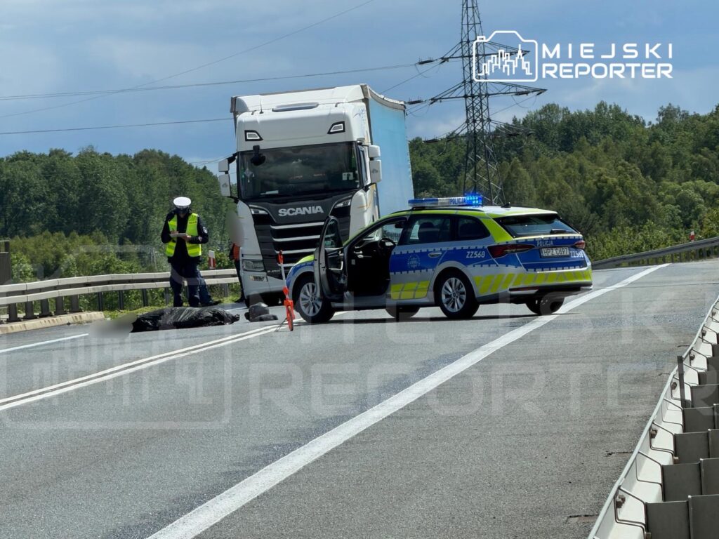 Policjant w odblaskowej kamizelce bada miejsce wypadku drogowego obok ciężarówki, na drodze z oznaczeniem policyjnym.