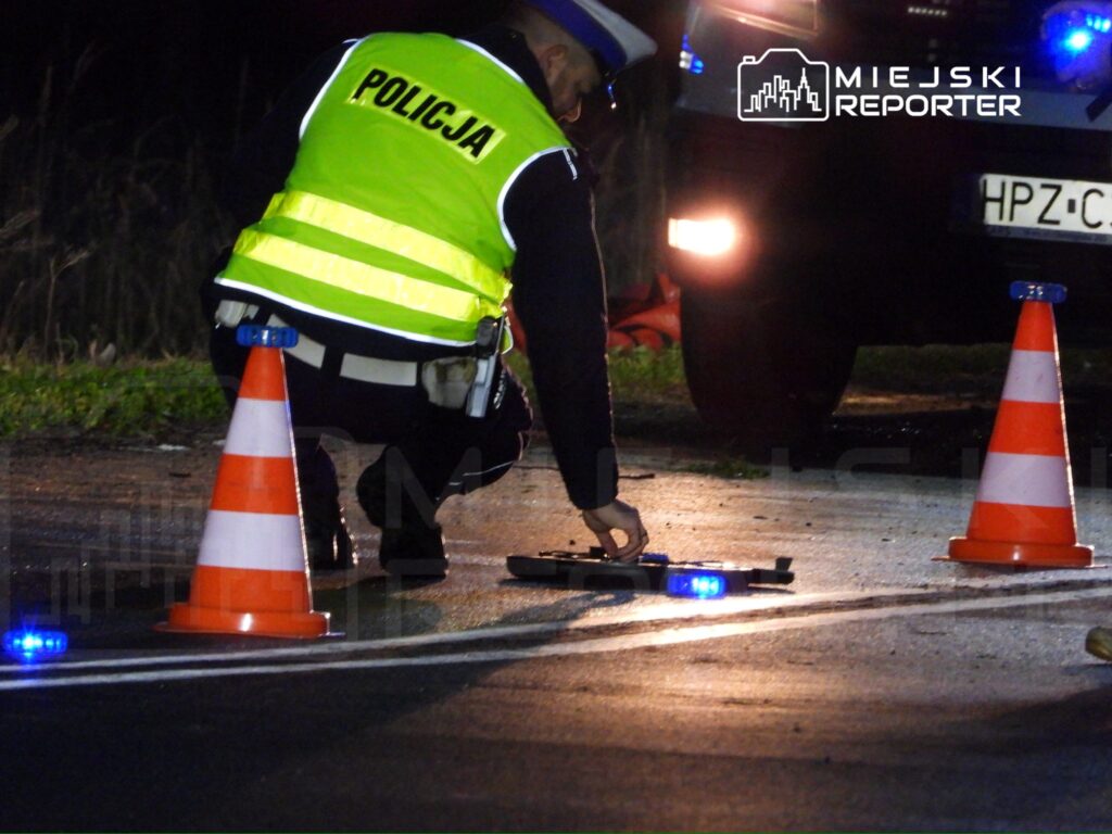 Policjant w odblaskowej kamizelce bada miejsce zdarzenia drogowego, obok pomarańczowe pachołki i policyjny radiowóz.