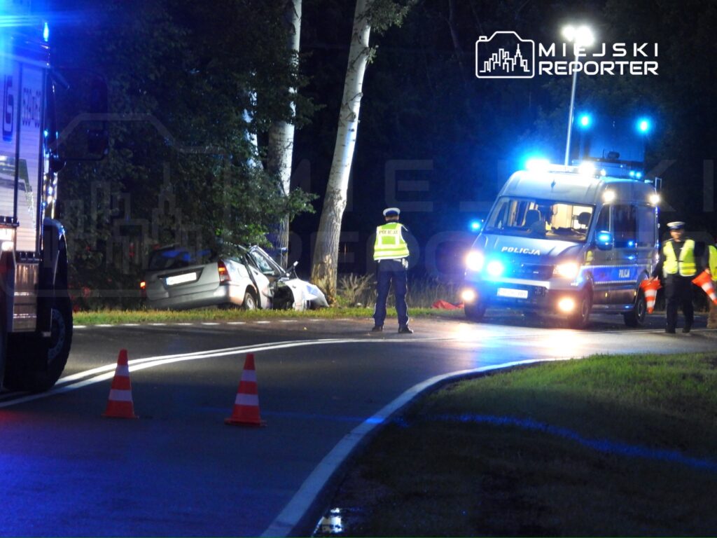 W nocy, na drodze, policjanci zabezpieczają miejsce wypadku samochodowego, obok stoi uszkodzony pojazd.